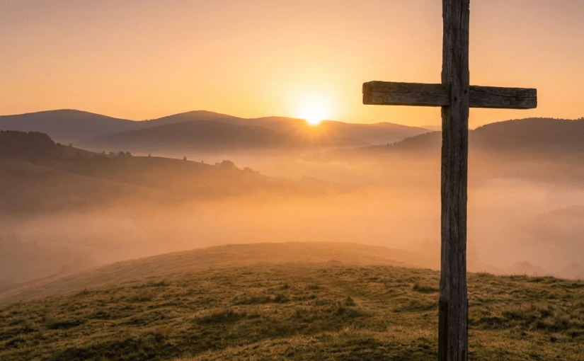 A wooden cross on a grassy hill with the sun rising over misty mountains.
